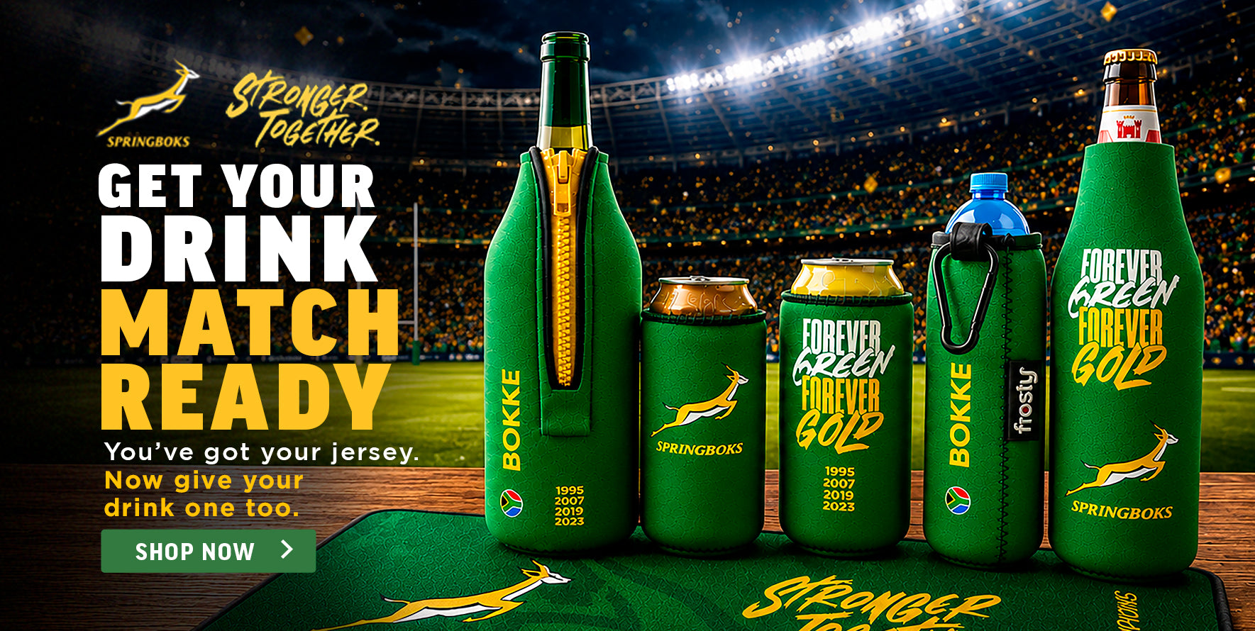 A promotional display of Springboks-branded drink containers on a green football field at night under stadium lights. The products include a large bottle, a can, a tumbler, and two small bottles, all wrapped in green Springboks-themed koozie-style sleeves with gold and white branding.